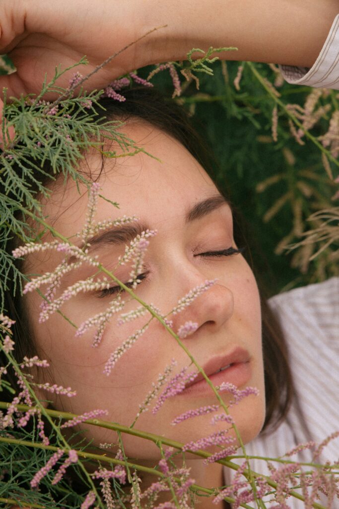 vrouw ligt met haar ogen dicht in het gras met plantjes voor haar gezicht.